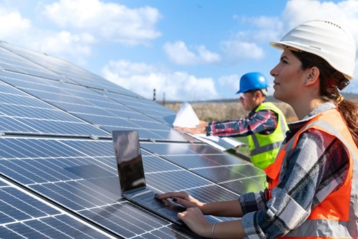 eine Frau und ein Mann mit Warnweste, Helm und Notebook an einem Dach mit Solarpanelen